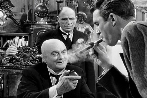A black and white photo of a man puffing on a cigar with two older men in suits standing before him.